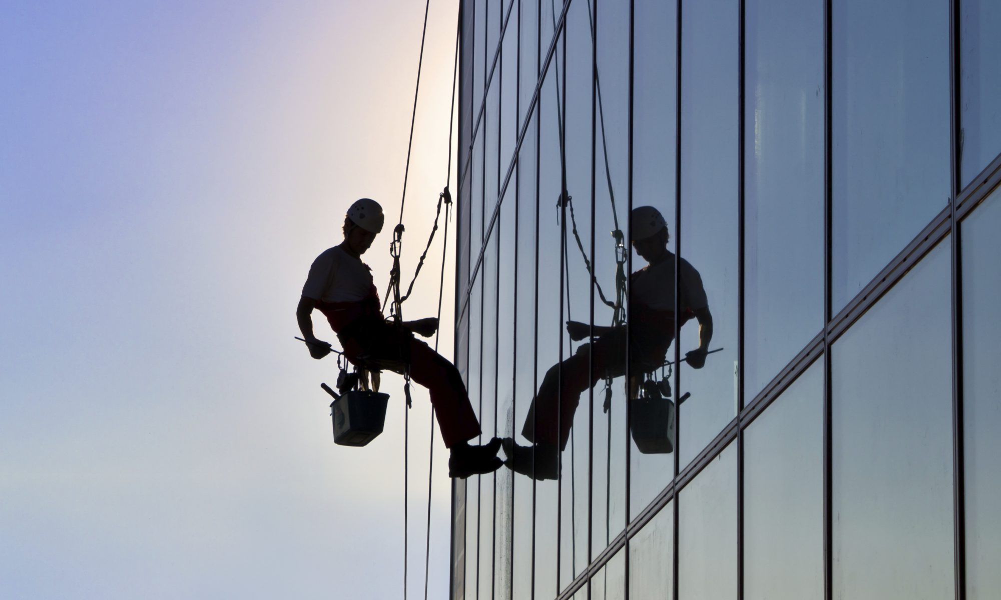 A rope access technician's sihlouette can been seen hanging off the side of a highrise building as he prepares his squeegee to wash and clean the windows in front of him.
